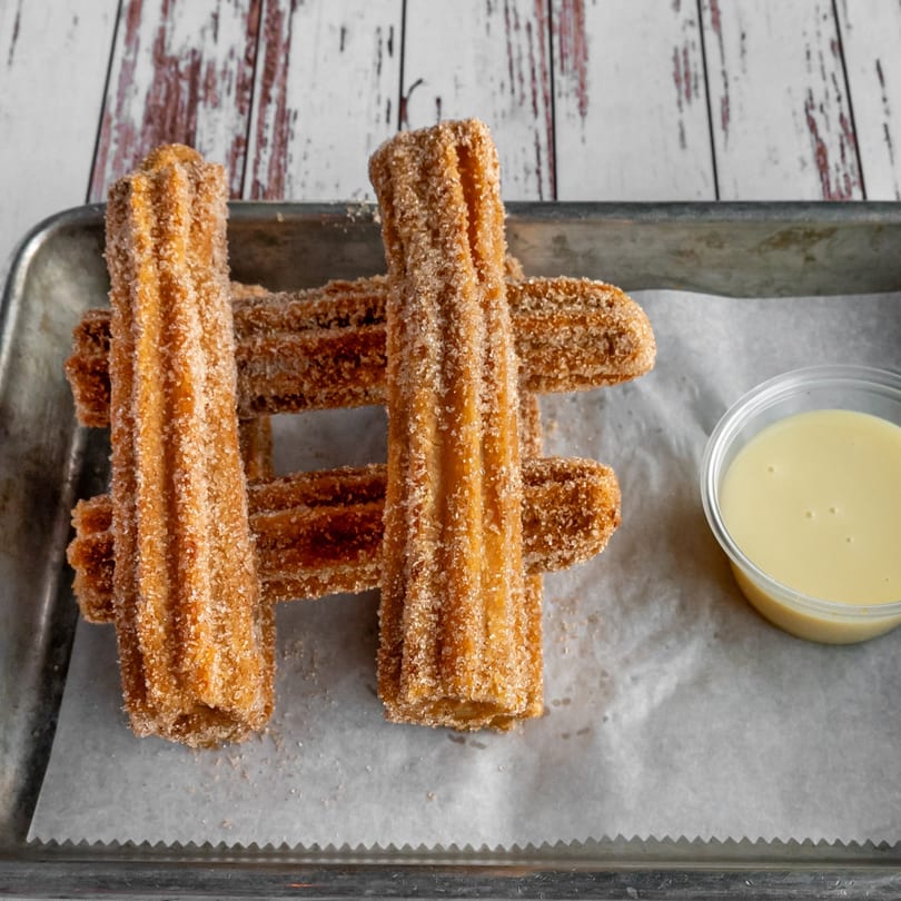 Safi Mediterranean Grill churros, coated in cinnamon sugar, with a side of dipping sauce on a metal tray.